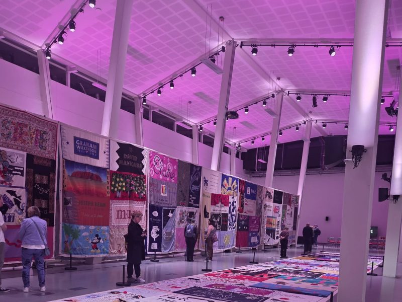 UK AIDS Memorial quilt laid out and displayed on the floor and walls, purple lighting, a few people walk by the quilts